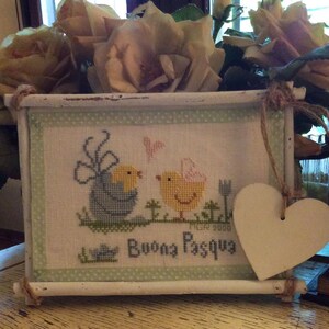 May include: A white wooden frame with a cross-stitch design of two yellow chicks with a blue chick in the background. The frame has a green and white polka dot border and a white heart-shaped tag hanging from the top. The cross-stitch design says "Buona Pasqua" which is Italian for "Happy Easter".