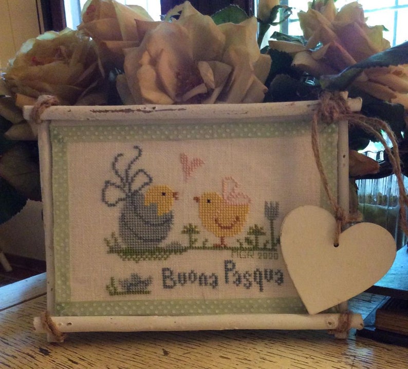 May include: A white wooden frame with a cross-stitch design of two yellow chicks with a blue chick in the background. The frame has a green and white polka dot border and a white heart-shaped tag hanging from the top. The cross-stitch design says "Buona Pasqua" which is Italian for "Happy Easter".