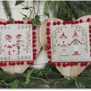Puede incluir: Dos placas de madera en forma de corazón con escenas navideñas bordadas. Cada una presenta un tejido cuadrado blanquecino con un diseño de punto de cruz de elfos en rojo y verde. Pompones rojos adornan los bordes. Las placas están rodeadas de vegetación.
