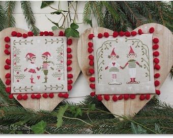 May include: Two heart-shaped wooden plaques with embroidered Christmas scenes. Each features a square, off-white fabric with a cross-stitched design of elves in red and green. Red pom-poms trim the edges. The plaques are surrounded by greenery.