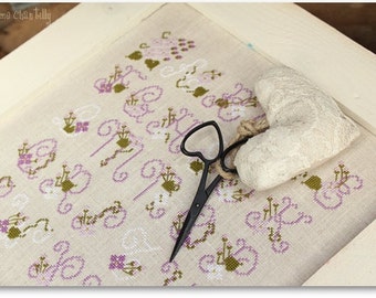 May include: A white linen fabric with a cross-stitch pattern of purple and green flowers and swirls. A white heart-shaped pillow and black scissors are on top of the fabric.