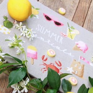 Könnte beinhalten: Ein Kreuzstich-Design auf einem grauen Stoff mit sommerlichen Essentials, darunter eine Zitrone, Sonnenbrillen, ein Glas Limonade, Eis, ein Krebs, eine Sandburg und der Text "Summer Essentials".
