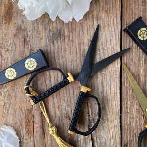 May include: Black and gold scissors with a tassel, a black and gold case, and a gold and black case on a wooden surface.