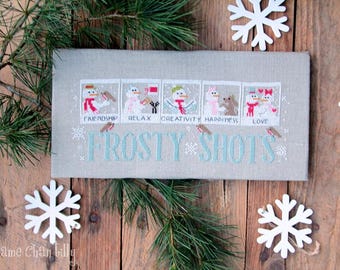 May include: A cross-stitch pattern featuring six different snowman designs, each with a unique personality. The designs are framed in a light brown linen fabric with the words "Frosty Shots" in a light blue font.