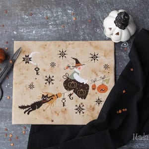 Puede incluir: Un diseño de punto de cruz que presenta una bruja en una escoba con un gato negro, calabazas y estrellas. El diseño está sobre una tela beige con una estética vintage. Una pequeña calabaza naranja y unas tijeras negras también están en la imagen.