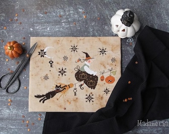 May include: A cross-stitch design featuring a witch on a broom with a black cat, pumpkins, and stars. The design is on a beige fabric with a vintage aesthetic. A small orange pumpkin and black scissors are also in the image.