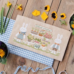 May include: A rustic wooden table displays a cross-stitch piece with two bunnies at a spring market. Pansies and daffodils in pots and a blue gingham cloth add color. The text "Spring Market" is visible.