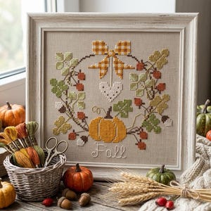 May include: A framed cross-stitch embroidery featuring a wreath of autumn leaves, berries, a checkered bow, a heart, and a pumpkin with the word Fall stitched below. A wicker basket holds colorful embroidery floss next to small pumpkins and wheat.
