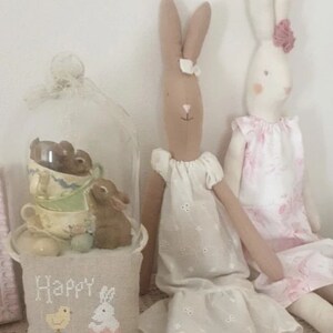 May include: Two handmade bunny dolls dressed in white and pink dresses. The dolls are sitting on a white crocheted surface with a pillow that says "Happy Easter" in front of them.