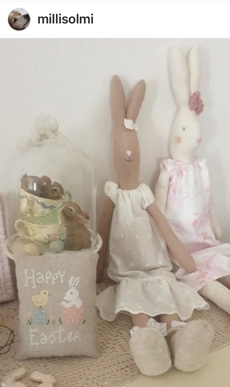 May include: Two handmade bunny dolls dressed in white and pink dresses. The dolls are sitting on a white crocheted surface with a pillow that says "Happy Easter" in front of them.