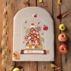 May include: A cross-stitch design depicting a village scene in autumnal colours, with pumpkins and apples. The word "Autumns" is stitched on a banner. The design is on a beige fabric, surrounded by apples, leaves, and ribbon on a wooden surface.