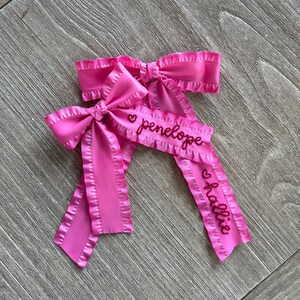 May include: Two pink satin hair bows with ruffled edges. The bows are personalized with the names "penelope" and "allie" in red embroidery.