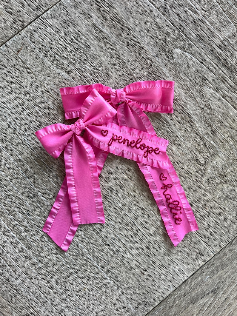 May include: Two pink satin hair bows with ruffled edges. The bows are personalized with the names "penelope" and "allie" in red embroidery.