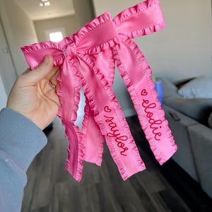 May include: Pink satin ribbon bow with ruffled edges and the names "Elodie" and "Taylor" embroidered in red.