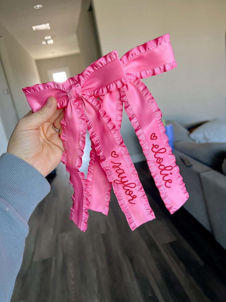 May include: Pink satin ribbon bow with ruffled edges and the names "Elodie" and "Taylor" embroidered in red.