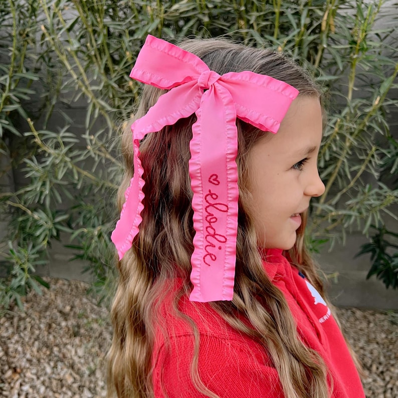 May include: Pink hair bow with a ruffled edge and a personalized name, "Elodie", embroidered in pink on the ribbon.