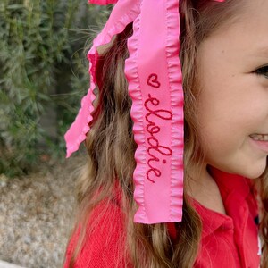 May include: Pink ruffled ribbon hair bow with the name "Elodie" embroidered in red thread.