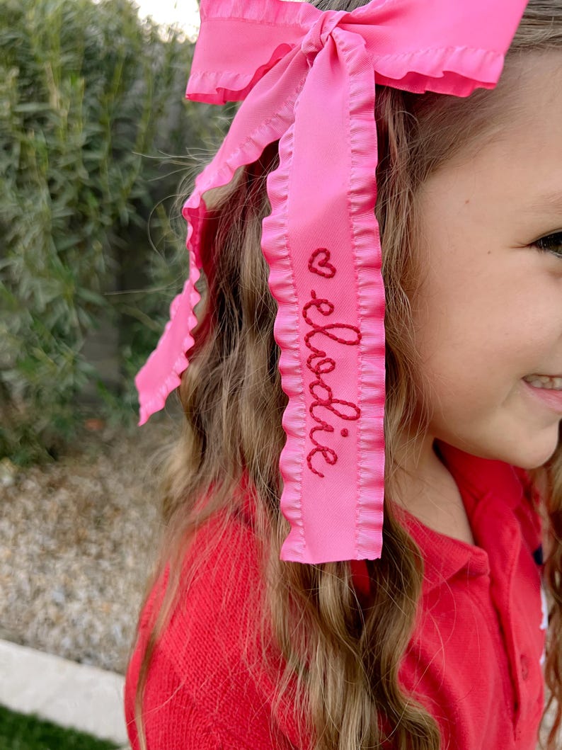 May include: Pink ruffled ribbon hair bow with the name "Elodie" embroidered in red thread.