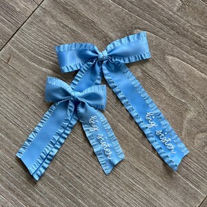 May include: Two blue satin hair bows with ruffled edges. The larger bow has the text "big sister" embroidered on it. The smaller bow has the text "little sister" embroidered on it.