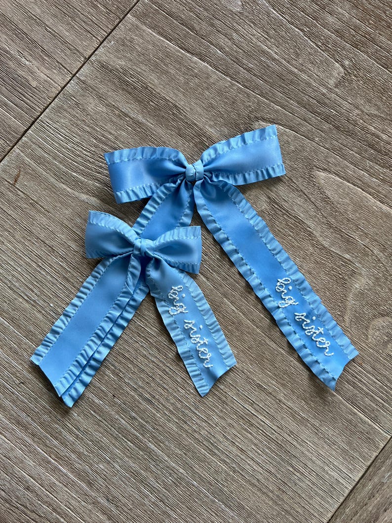 May include: Two blue satin hair bows with ruffled edges. The larger bow has the text "big sister" embroidered on it. The smaller bow has the text "little sister" embroidered on it.