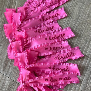 May include: Pink satin ribbon bows with ruffled edges and embroidered names, including Hayden, Reagan, Scottlyn, Harper, Kinley, Kelsey, Remi, Brielle, Hazel, and Liv Mae.