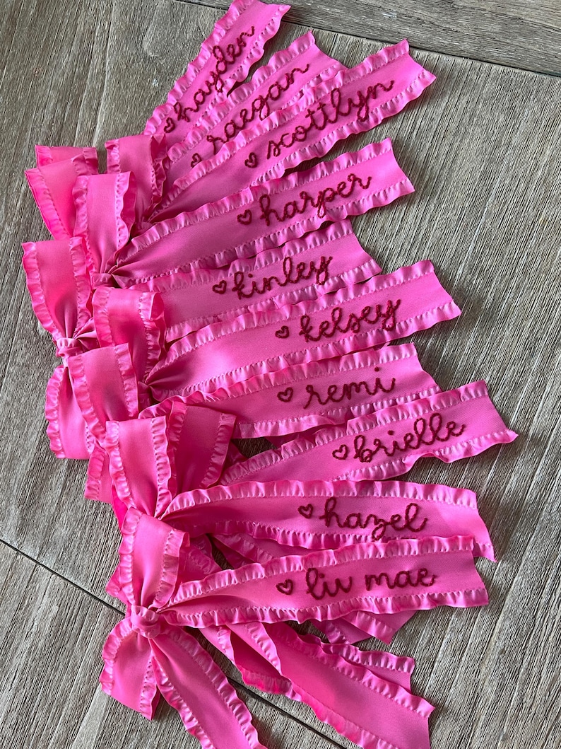May include: Pink satin ribbon bows with ruffled edges and embroidered names, including Hayden, Reagan, Scottlyn, Harper, Kinley, Kelsey, Remi, Brielle, Hazel, and Liv Mae.