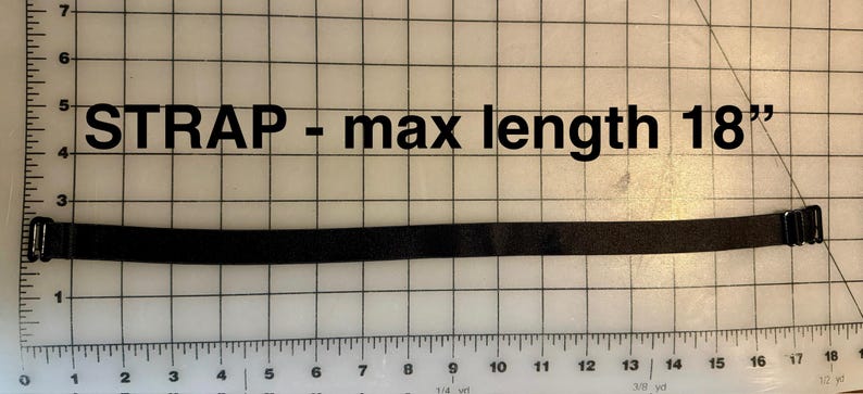 May include: A black strap with metal hardware, measuring a maximum length of 18 inches. The text "STRAP - max length 18" is printed above the strap. The strap is placed on a grid background.