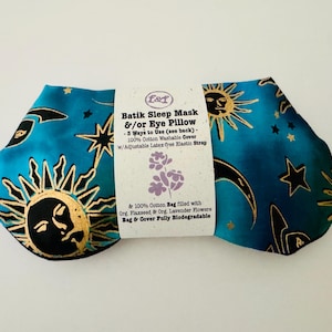 May include: A batik sleep mask and eye pillow with a teal background and gold sun, moon, and star designs. The mask has a white label with text describing its features, including a washable cotton cover and adjustable strap.