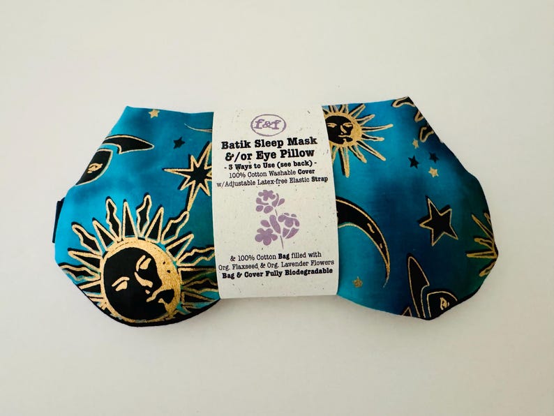 May include: A batik sleep mask and eye pillow with a teal background and gold sun, moon, and star designs. The mask has a white label with text describing its features, including a washable cotton cover and adjustable strap.