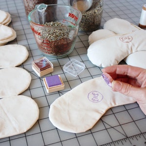 Puede incluir: Primer plano de una persona estampando una m&aacute;scara de ojos blanca con una almohadilla de tinta morada. La m&aacute;scara de ojos tiene la palabra "Feet" estampada. Hay varias otras m&aacute;scaras de ojos en el fondo, junto con una taza medidora llena de lavanda seca y un peque&ntilde;o recipiente de tinta.