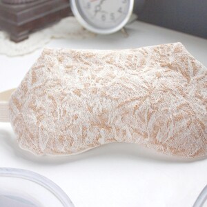 May include: A beige eye mask with a white floral pattern. The mask has an elastic strap for a comfortable fit.