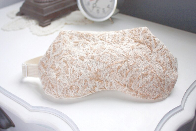 May include: A beige eye mask with a white floral pattern. The mask has an elastic strap for a comfortable fit.