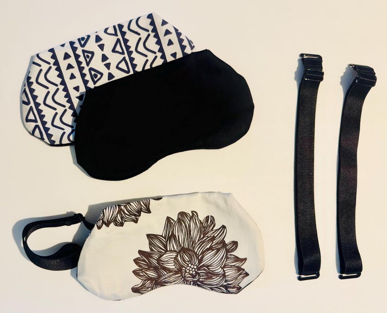 May include: Two sleep masks, one black and one with a white and navy blue geometric pattern, are displayed. A third mask features a white background with a brown floral design. Two black elastic straps with metal adjusters are also included.