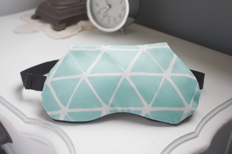 May include: A light blue sleep mask with a white geometric pattern. The mask has an adjustable black elastic strap.