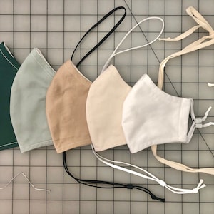 May include: Five fabric face masks with different colored ties. The masks are in shades of green, light green, beige, cream, and white. The masks are laid out in a row, with the ties hanging down.