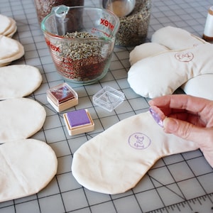 May include: A close-up of a person stamping a white eye mask with a purple ink pad. The eye mask has the word "Feet" stamped on it. There are several other eye masks in the background, along with a measuring cup filled with dried lavender and a small jar of ink.
