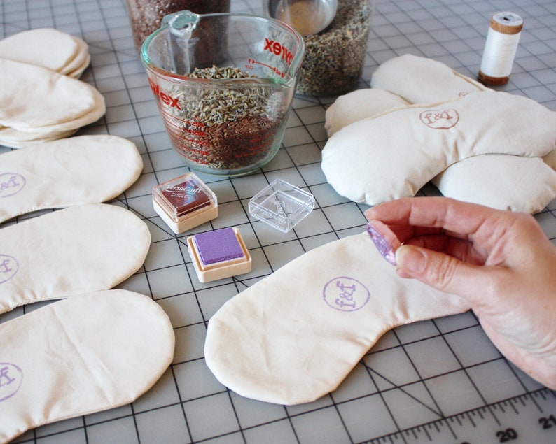 Puede incluir: Una configuraci&oacute;n artesanal con varias m&aacute;scaras para los ojos de color blanquecino con un logotipo estampado, junto a recipientes de lavanda seca y semillas de lino. Se muestra una mano estampando una m&aacute;scara con una almohadilla de tinta p&uacute;rpura. La escena sugiere productos de aromaterapia hechos a mano.