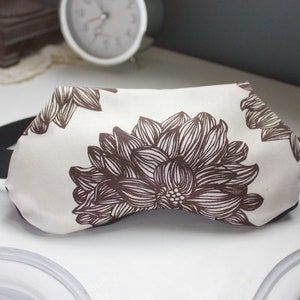May include: A white sleep mask with a brown floral pattern. The mask has an adjustable strap.