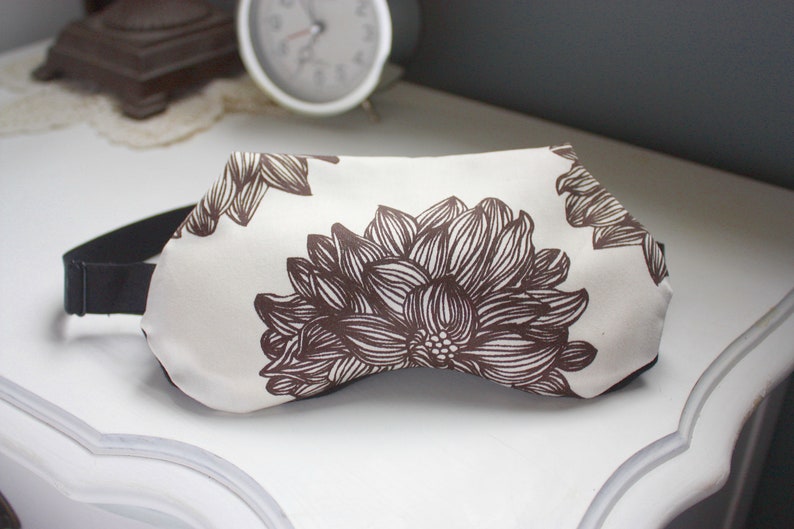 May include: A white sleep mask with a brown floral pattern. The mask has an adjustable strap.