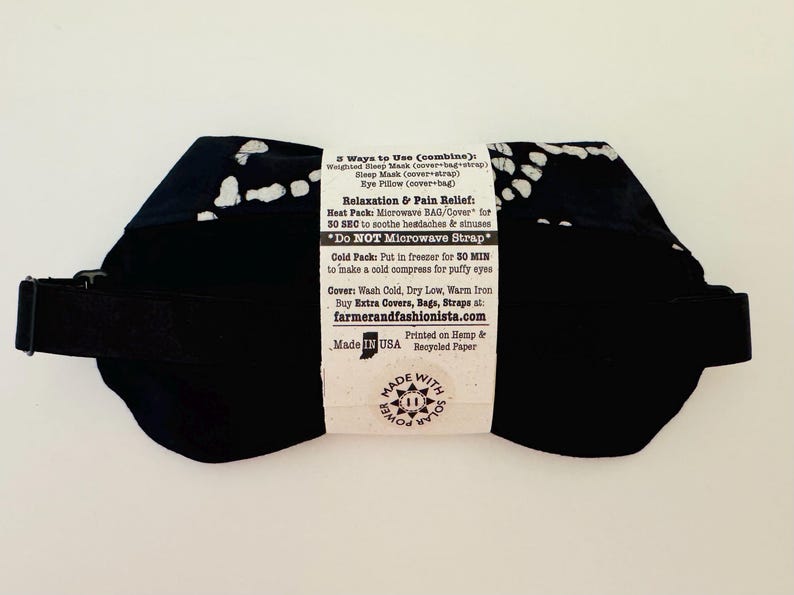 May include: Black sleep mask with white spotted pattern and an adjustable strap. The mask has a label with instructions for use, including microwave and cold pack options. Made in the USA.