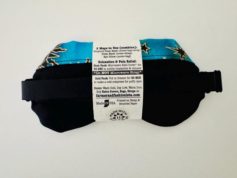 May include: A black eye mask with a turquoise and gold star pattern on the top. The mask has a black strap and a white label with text about its uses and care instructions. The product is made in the USA.