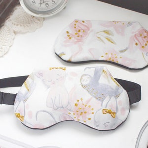 K&ouml;nnte beinhalten: Zwei Schlafmasken mit schwarzen Gummib&auml;ndern. Eine Maske hat ein pinkfarbenes und goldenes Blumenmuster. Die andere Maske hat einen wei&szlig;en Hintergrund mit einer pinkfarbenen Katze, einem grauen Faultier und einem pinkfarbenen Hasen.