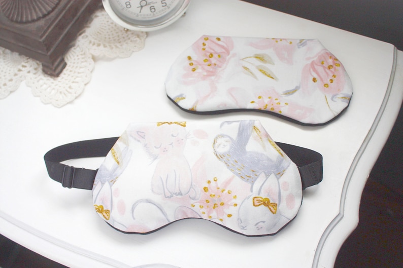 K&ouml;nnte beinhalten: Zwei Schlafmasken mit schwarzen Gummib&auml;ndern. Eine Maske hat ein pinkfarbenes und goldenes Blumenmuster. Die andere Maske hat einen wei&szlig;en Hintergrund mit einer pinkfarbenen Katze, einem grauen Faultier und einem pinkfarbenen Hasen.