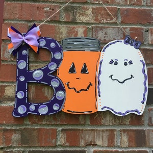 May include: A wooden Halloween sign spelling "BOO". The "B" is purple with bubble accents and a bow. The "O" is an orange jack-o'-lantern jar. The second "O" is a white ghost with a purple bow. The sign hangs from a string.
