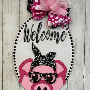 May include: A pink pig with black glasses and a black bandana with white polka dots. The pig is on a white oval wooden sign with a black polka dot border. The sign says "Welcome" in black letters with white rhinestones. A pink and white polka dot bow with a pink ribbon is on top of the sign.