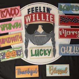 May include: A painted wooden sign featuring a portrait of Willie Nelson with sunglasses and braids, surrounded by rectangular signs with words like "Loved," "Springy," and "Baked." The signs are in various colors.