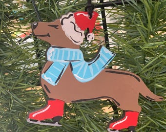 May include: A brown dachshund ornament wearing a red Santa hat, a light blue striped scarf, and red ice skates. The flat, painted wooden cutout is suspended by a black string, depicting the dog in a side profile view with a cheerful expression.