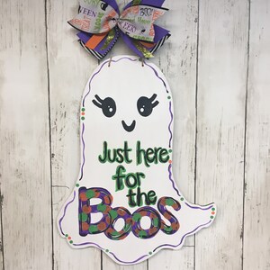 May include: A white ghost door hanger with a purple, green, and orange bow. The ghost has a smiling face and the text "Just here for the boos" in green, orange, and purple.