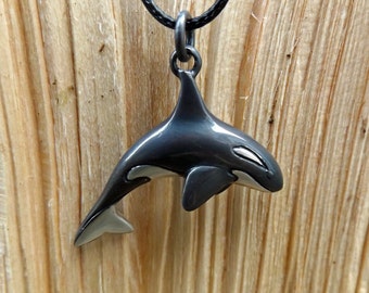 May include: A silver-toned orca pendant on a black cord. The pendant features a detailed design of a killer whale with black and white coloring. The orca is suspended from a circular bail, and the cord is knotted at the top.