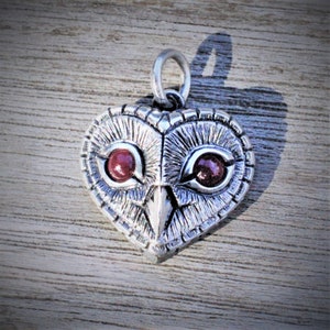 May include: A silver-colored owl pendant with a heart-shaped design. The pendant features a detailed owl face with red gemstone eyes and a textured surface. The pendant has a loop at the top for a chain.